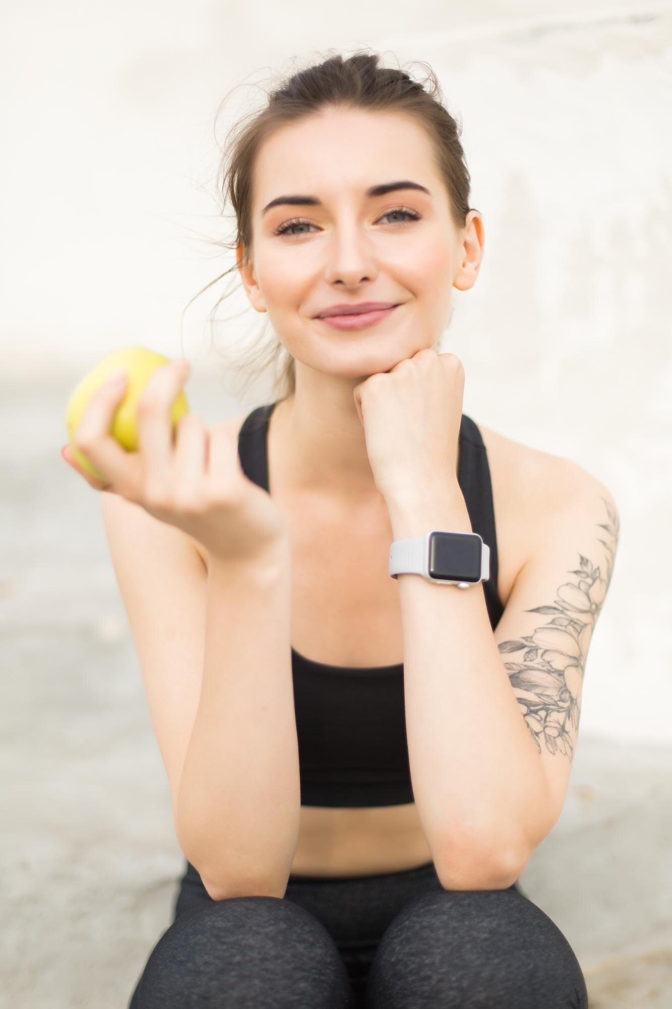 Mulher jovem satisfeita segurando uma fruta com uma mão e usando um smartwatch com roupas de treino, tatuagem no braço, tatuagem de flores, tatuagem feminina, smartwatch bem avaliado, look fitness, garota jovem sexy