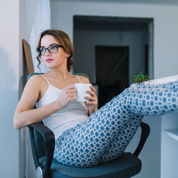 Mulher de pijama no home office, tomando café, sentada na cadeira com os pés sobre a mesa de trabalho.