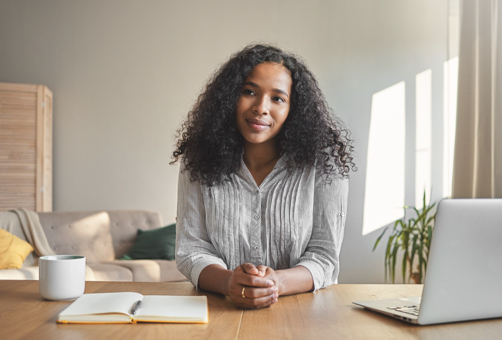 Mulher preta bela em home office, debruçada sobre a mesa de trabalho refletindo.
